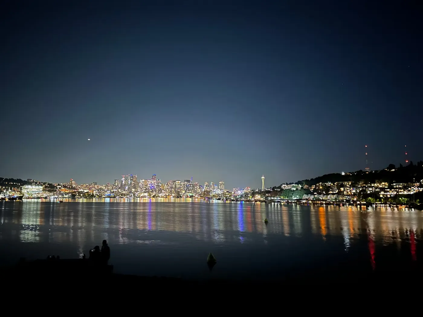 A city skyline reflected across dark water at night, the Space Needle lit at the right of the frame.