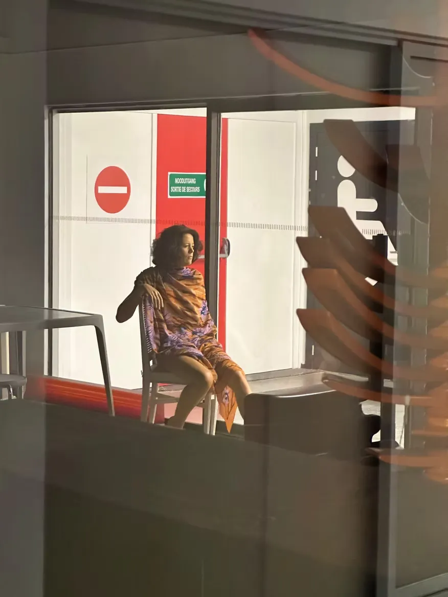 A seated figure framed by a doorway, seen through a window with warm reflections across the glass.