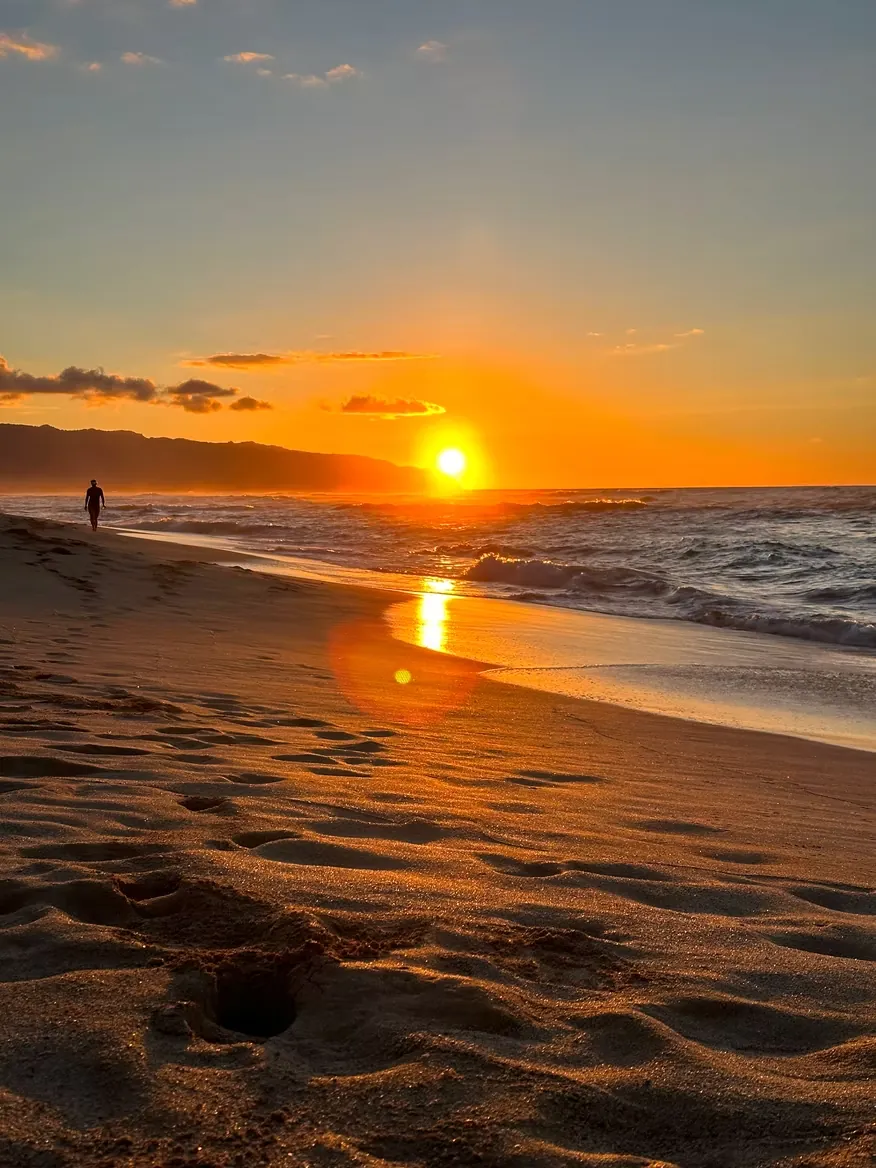 A low sun burning orange over breaking surf, a lone walker on the wet sand.