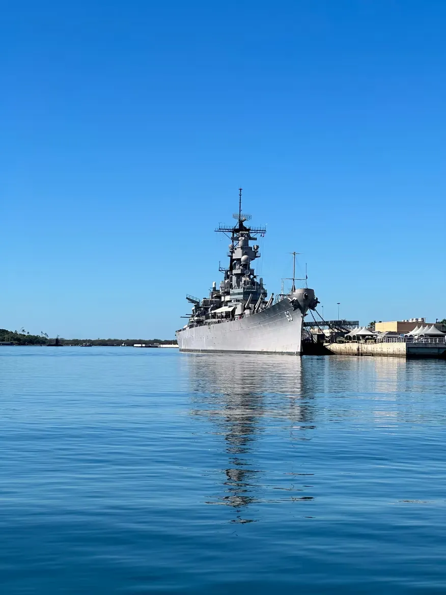 A grey battleship moored under a clear blue sky, mirrored in still harbour water.