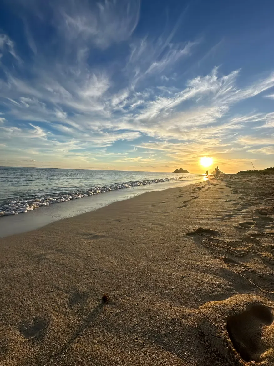 Wide cirrus clouds over a beach at sunset, two small figures at the waterline near offshore islands.